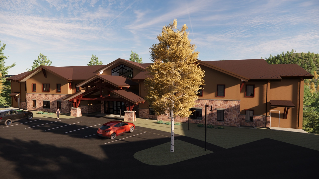 A modern lodge-style building with a stone and stucco exterior, featuring a covered entrance with wooden beams. The surrounding landscape includes trees, greenery, and a parking lot with a few cars. The structure blends into the natural setting, creating a warm and inviting atmosphere.
