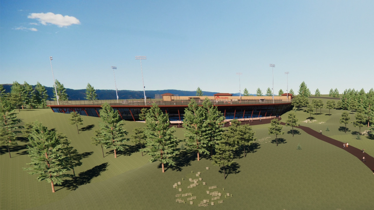 A stadium-style sports facility built into a hillside, surrounded by trees and open landscape. Tall floodlights line the field, and a walking path leads to the entrance, blending the venue into its natural setting.