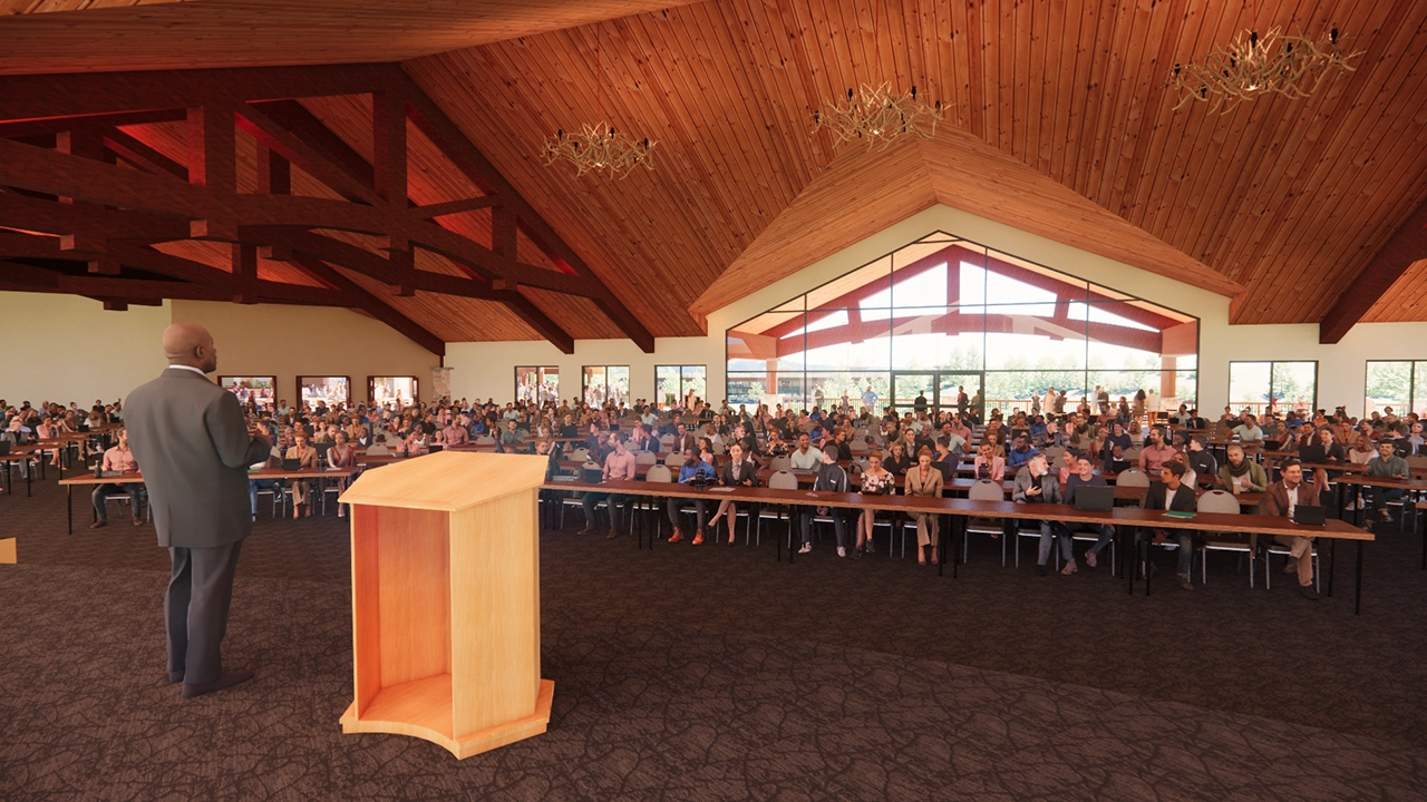 A spacious conference hall with high wooden ceilings, large windows, and chandeliers, filled with attendees seated at long tables. A speaker stands at the podium, addressing the audience, many of whom are taking notes or using laptops. The natural light and warm wood tones create an inviting and professional atmosphere.