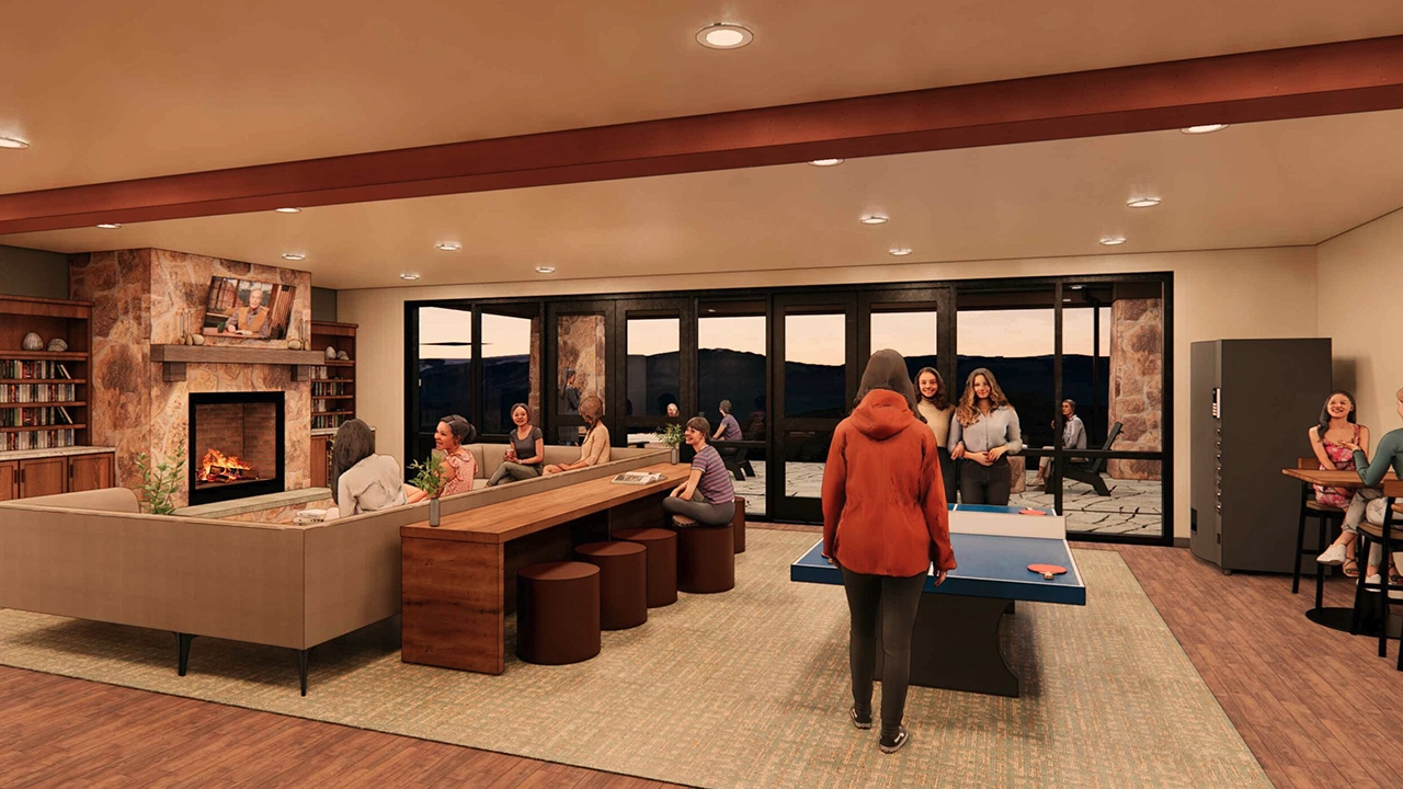 A warm and inviting lounge area featuring a stone fireplace, built-in bookshelves, and a large seating arrangement. The space includes a long communal table, a ping pong table, and a vending machine. People are socializing, playing games, and relaxing, while large glass doors offer a scenic view of the sunset and an outdoor patio.
