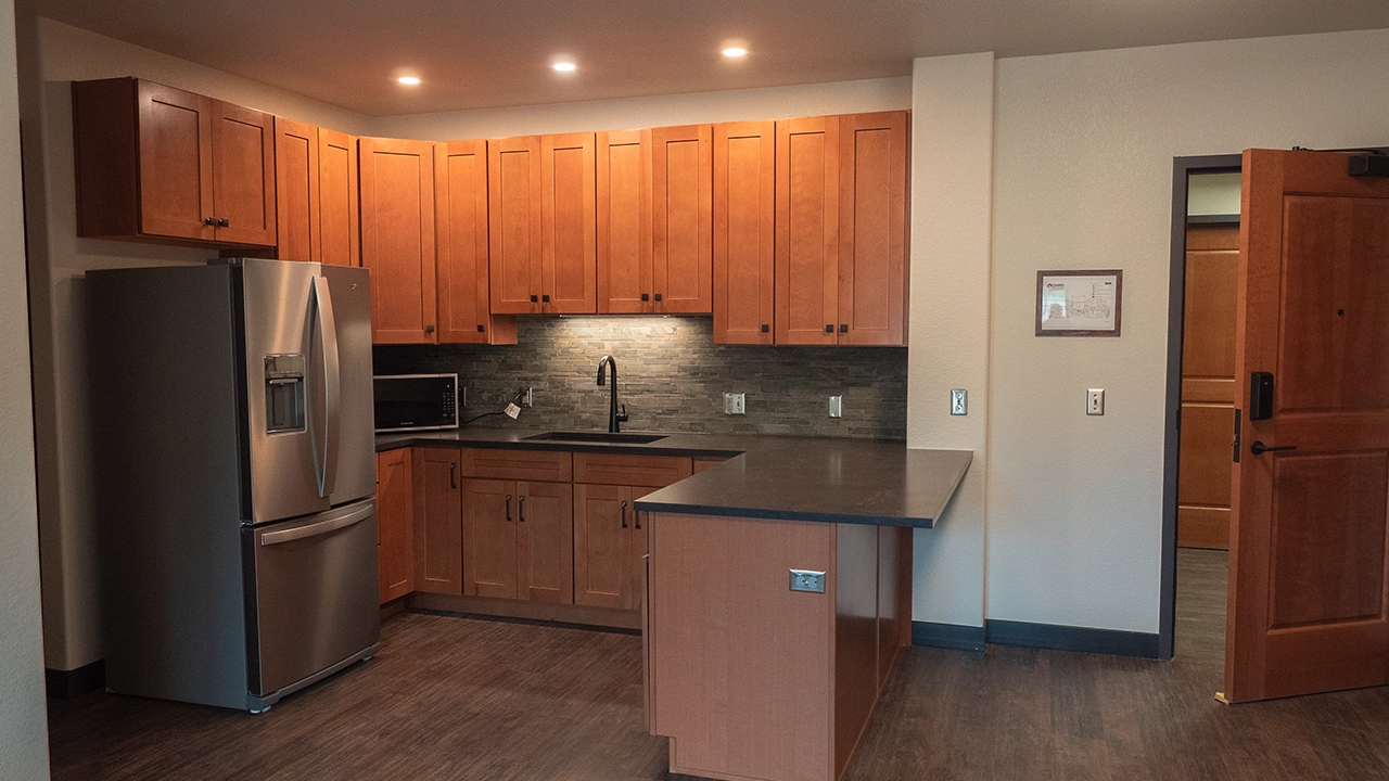 A modern kitchen with warm wooden cabinetry, a sleek black countertop, and a stylish backsplash. It features stainless steel appliances, including a large refrigerator and a microwave. Recessed lighting provides a cozy ambiance, while a small peninsula adds counter space and functionality.