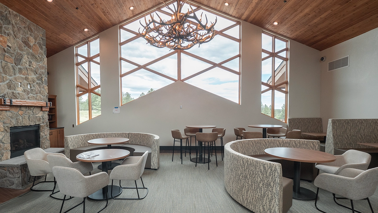 A cozy lounge area with modern and rustic elements, featuring a stone fireplace, high wooden ceilings, and large geometric windows that let in natural light. The space is furnished with round tables, plush chairs, and curved booths, while an antler chandelier adds a distinctive mountain-lodge aesthetic.
