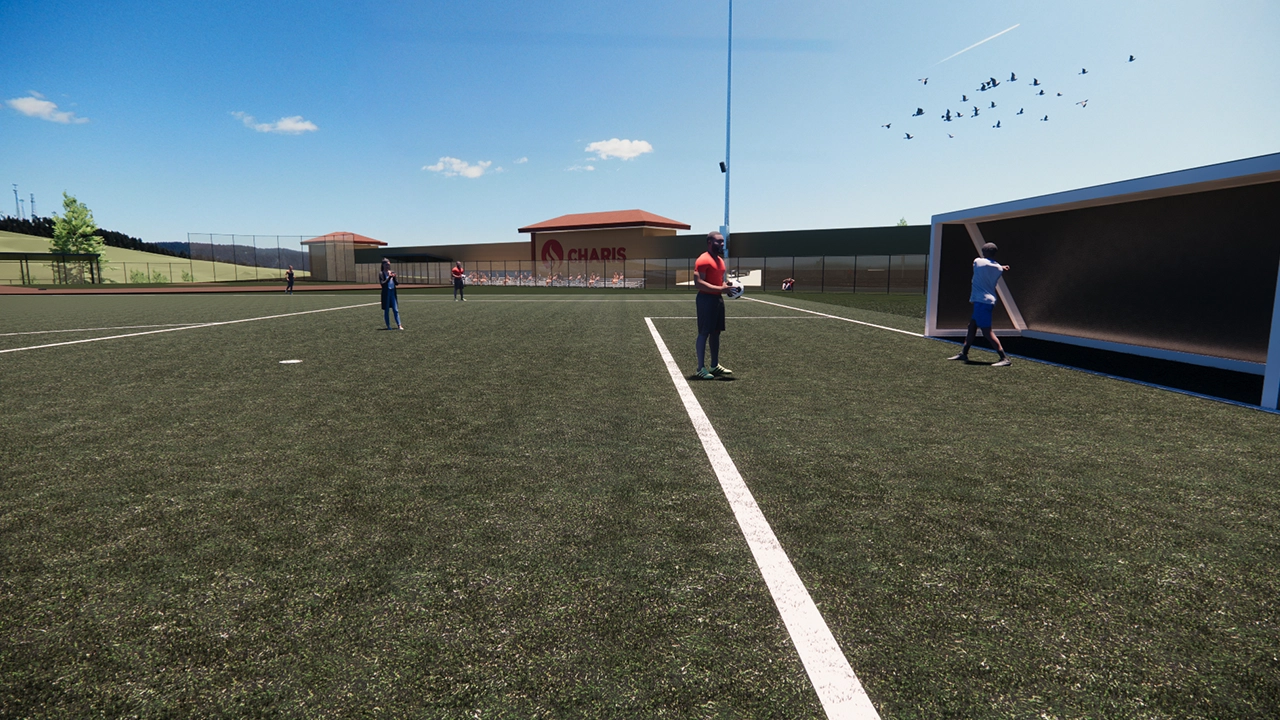 An outdoor sports field with artificial turf, where several people are engaged in activities under a clear blue sky. A player in a red shirt stands near the sideline, while another practices against a rebound wall. In the background, a building with the "Charis" logo is visible, along with a group of people near the fence and birds flying overhead.